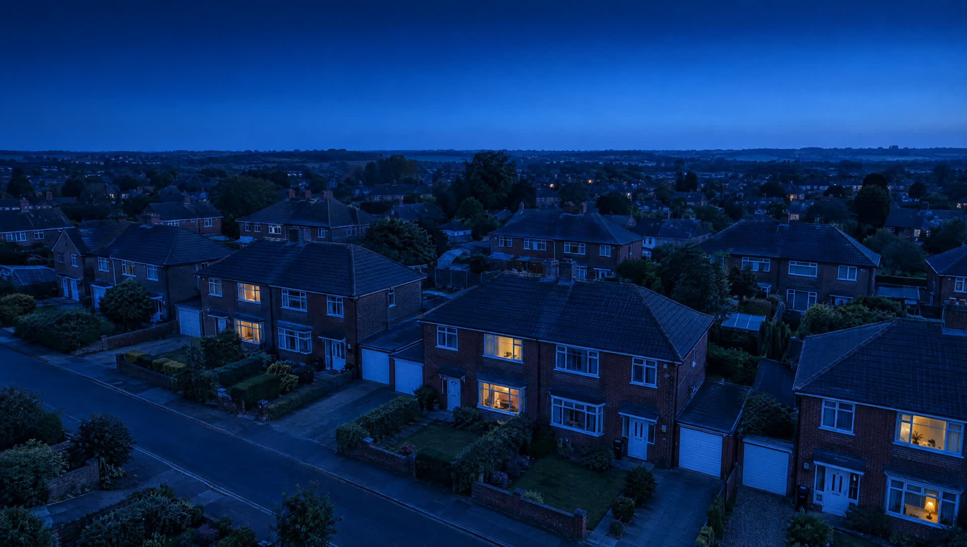 Evening aerial view of a British suburban street, warm light in the windows of the family homes